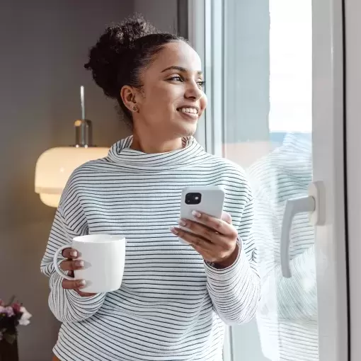 Happy woman standing in window of new phone holding drink and phone